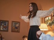 Young woman smiles and holds her arms out wide, looks as if she is playing charades with a group of people in a living room with Christmas decorations in the background.