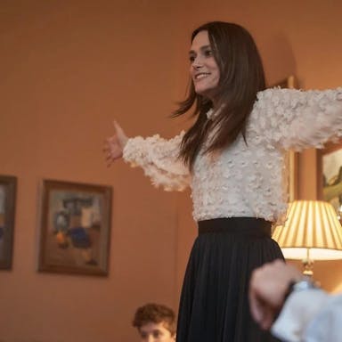 Young woman smiles and holds her arms out wide, looks as if she is playing charades with a group of people in a living room with Christmas decorations in the background. 