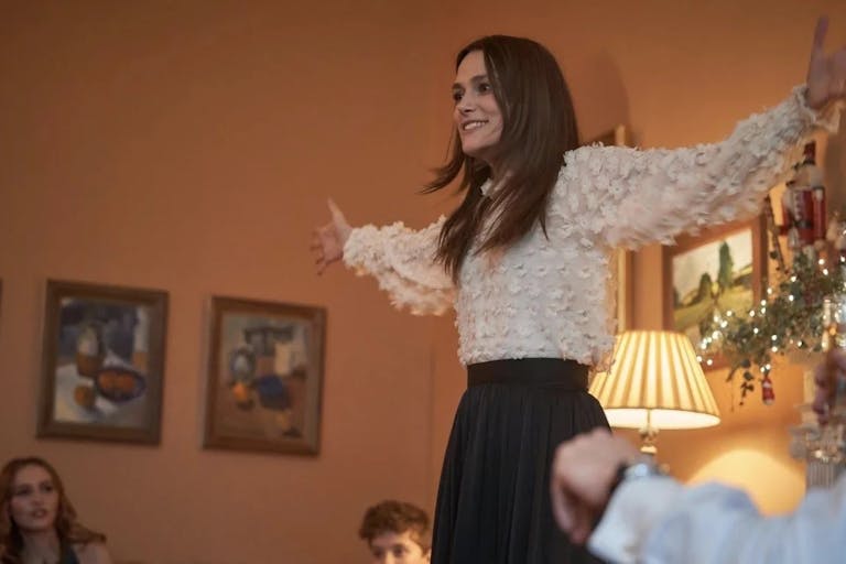 Young woman smiles and holds her arms out wide, looks as if she is playing charades with a group of people in a living room with Christmas decorations in the background. 