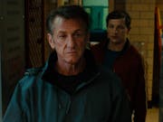 A tired looking middle aged white man in a paramedic locker room with a young white man standing behind him