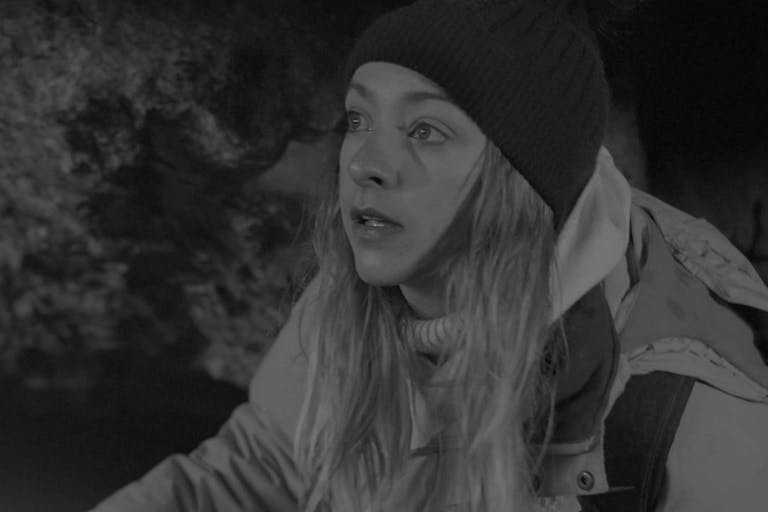 Black and white still of a white woman with long blonde hair wearing a wooly beanie hat, turtleneck jumper, and waterproof jacket