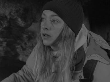 Black and white still of a white woman with long blonde hair wearing a wooly beanie hat, turtleneck jumper, and waterproof jacket
