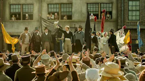 A dozen people standing on a stage wearing 19th Century attire, addressing a crowd of celebrating people