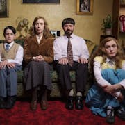 A white family of a Mother, Father, teen daughter and young son, wearing dated, 'respectable' clothing sitting on the sofa and floor