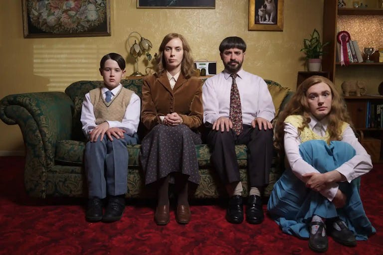 A white family of a Mother, Father, teen daughter and young son, wearing dated, 'respectable' clothing sitting on the sofa and floor