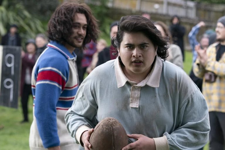 A young Maori man in a rugby shirt holding a battered rugby ball looking determined