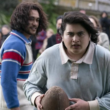 A young Maori man in a rugby shirt holding a battered rugby ball looking determined