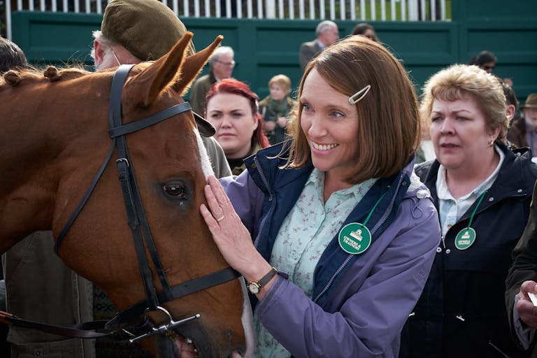 Smiling woman with a brown bob hairstyle puts her hand on a horse's head, she is wearing a green badge that indicates she is at a horse racecourse. 