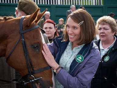 Smiling woman with a brown bob hairstyle puts her hand on a horse's head, she is wearing a green badge that indicates she is at a horse racecourse. 