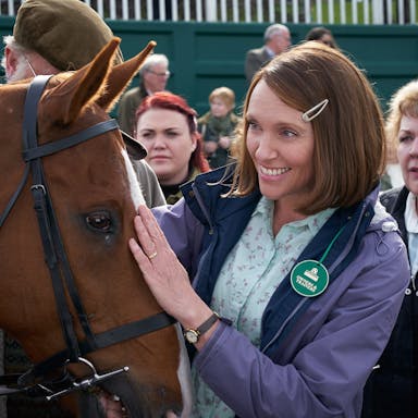 Smiling woman with a brown bob hairstyle puts her hand on a horse's head, she is wearing a green badge that indicates she is at a horse racecourse. 