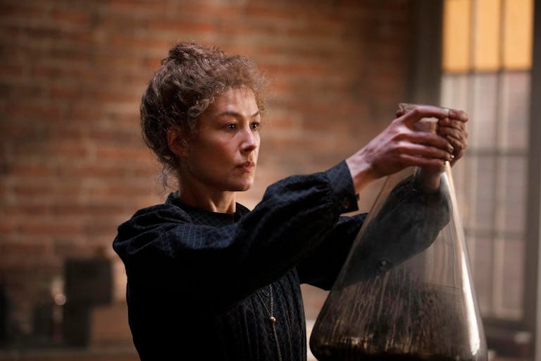A woman in period dress in a laboratory holding a very large beaker up