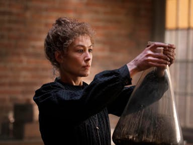 A woman in period dress in a laboratory holding a very large beaker up