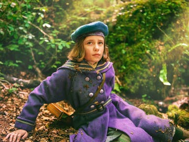 Young girl with brown hair sits on the floor of a woodland area, wearing an old-fashioned button-up purple coat and a blue hat