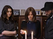 A woman wearing a brown wig, flanked by another brunette woman and a man in a black hat, lights two candles