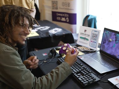 A young Black man smiling playing a video on a laptop using a purple controller