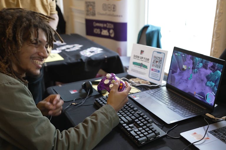 A young Black man smiling playing a video on a laptop using a purple controller