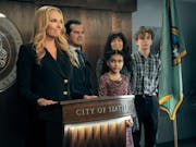 Blonde woman stands at a lectern that reads 'City of Seattle', surrounded by two adults and two children