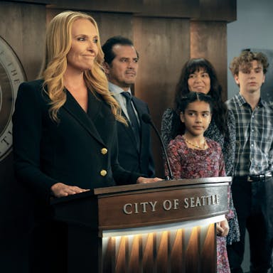 Blonde woman stands at a lectern that reads 'City of Seattle', surrounded by two adults and two children
