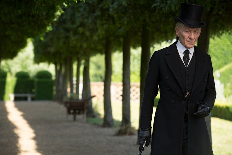 A tall man in long black coat and top hat, walking with a cane through a large stately garden between two lines of trees