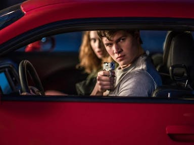 A young white man pointing a gun out of the window of a red car, a young blonde woman sits in the passenger seat