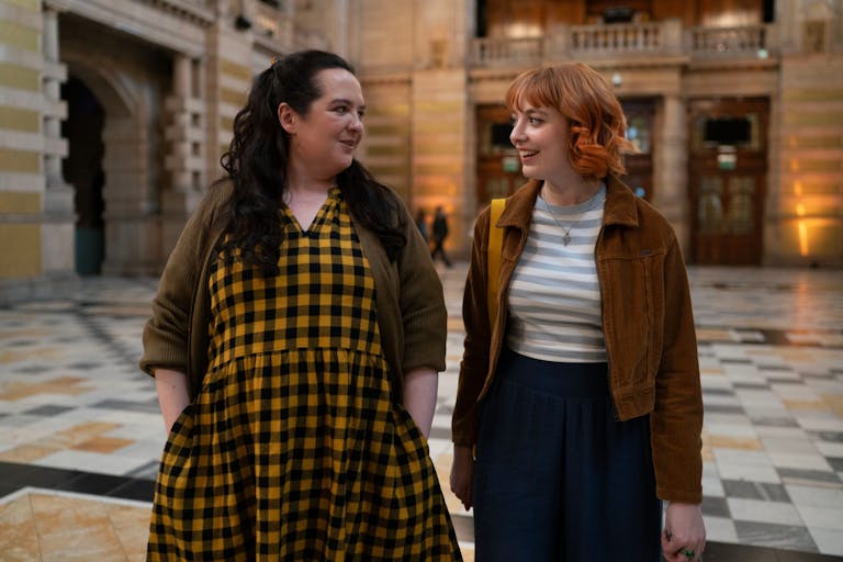 A young white woman with long black hair and yellow gingham dress and a young white woman in corduroy jacket walk together through a museum foyer smiling