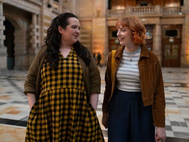 A young white woman with long black hair and yellow gingham dress and a young white woman in corduroy jacket walk together through a museum foyer smiling