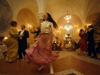 A young white woman with long dark hair in a frilly white blouse and pink flouncy skirt dancing with abandon in a grand banquet hall