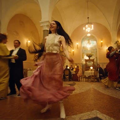 A young white woman with long dark hair in a frilly white blouse and pink flouncy skirt dancing with abandon in a grand banquet hall