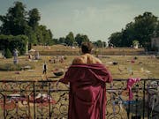 A young white man in a red dressing gown with his shoulders exposed, looking from a balcony at a large estate's garden strewn with the remnants of a wild party