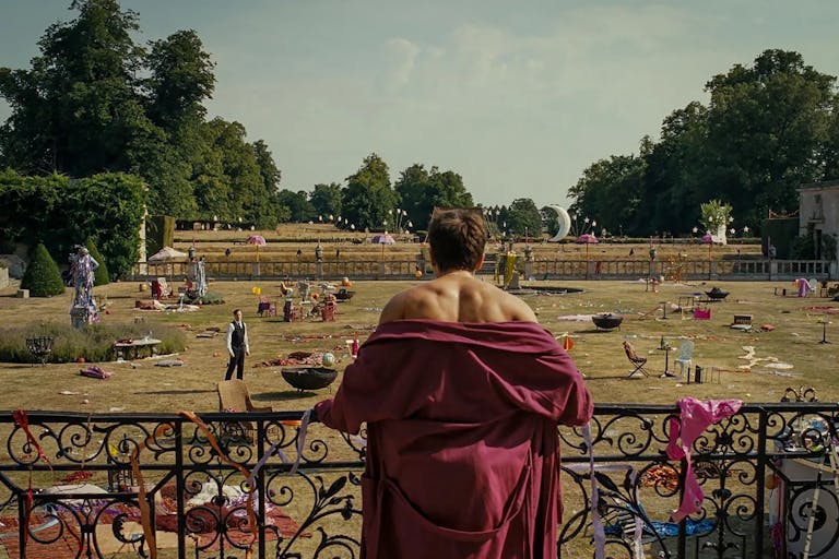 A young white man in a red dressing gown with his shoulders exposed, looking from a balcony at a large estate's garden strewn with the remnants of a wild party