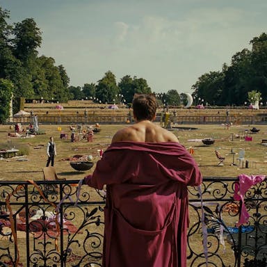 A young white man in a red dressing gown with his shoulders exposed, looking from a balcony at a large estate's garden strewn with the remnants of a wild party