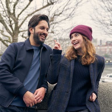 Young man and woman lean against a wall alongside a city river and smile at each other