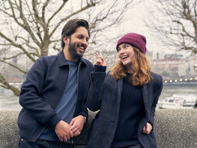 Young man and woman lean against a wall alongside a city river and smile at each other