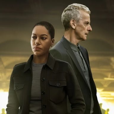 A young Black woman and an older white man stand looking serious in a dark setting 