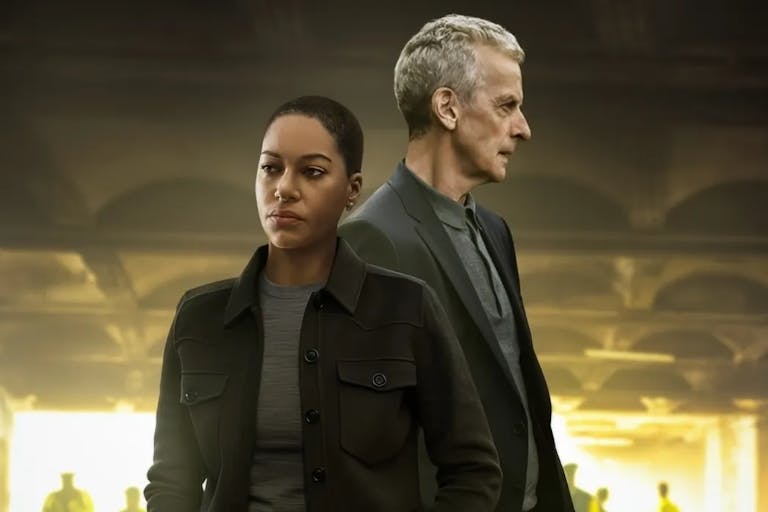 A young Black woman and an older white man stand looking serious in a dark setting