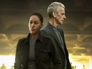 A young Black woman and an older white man stand looking serious in a dark setting