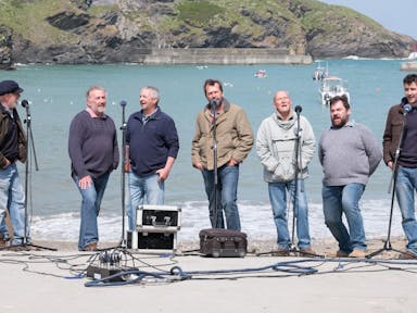 Seven men standing on a beachfront with the sea and cliffs behind them singing into mics