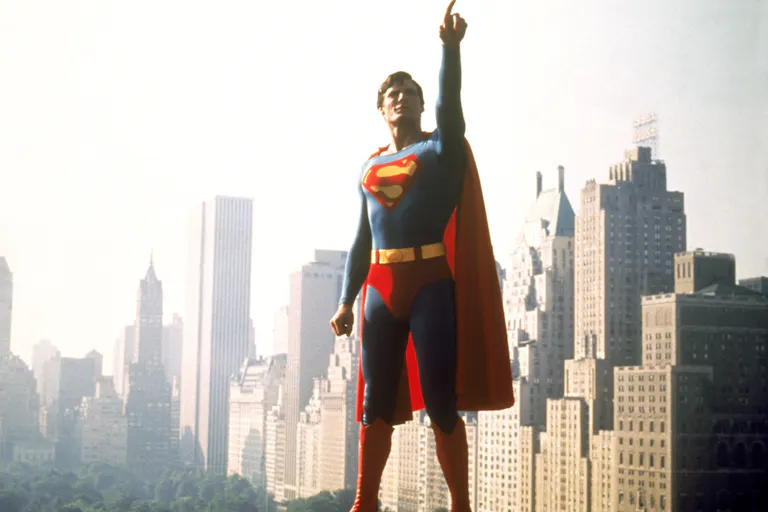 A white man in Superman costume, blue and red suit with red cape, pointing upwards, flying through New York