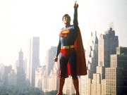 A white man in Superman costume, blue and red suit with red cape, pointing upwards, flying through New York