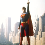 A white man in Superman costume, blue and red suit with red cape, pointing upwards, flying through New York