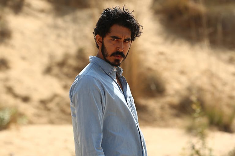 Bearded man in a blue shirt looks broodingly into the distance against a desert-like background.