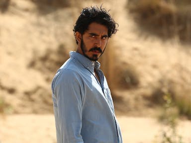 Bearded man in a blue shirt looks broodingly into the distance against a desert-like background. 