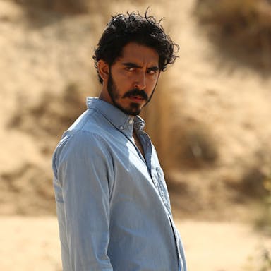 Bearded man in a blue shirt looks broodingly into the distance against a desert-like background. 