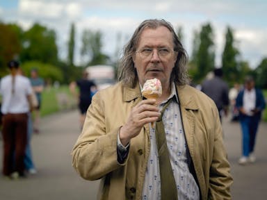An older bedraggled looking white man with loose coat, shirt and tie eating a melting ice cream while walking through a park