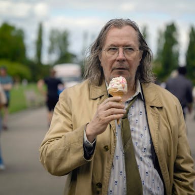 An older bedraggled looking white man with loose coat, shirt and tie eating a melting ice cream while walking through a park