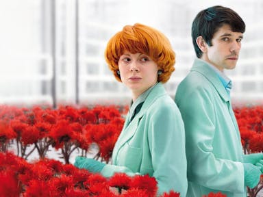 A couple stand back to back in green lab coats and gloves surrounded by red flowers.