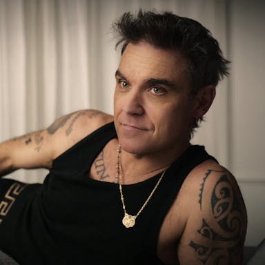 A middle aged white man with dark hair and tattoos, lounging in black vest and boxers, wearing a small gold tiger chain necklace