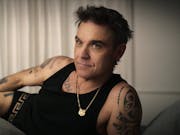 A middle aged white man with dark hair and tattoos, lounging in black vest and boxers, wearing a small gold tiger chain necklace