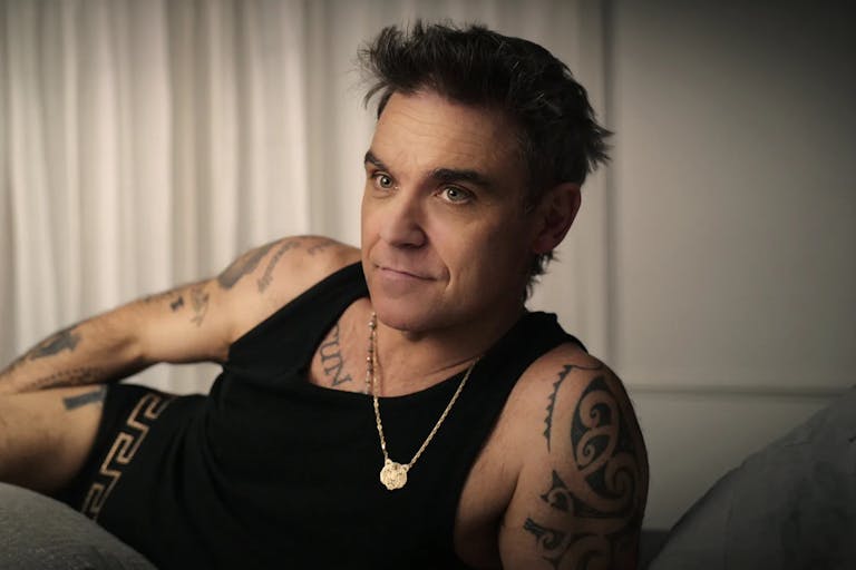 A middle aged white man with dark hair and tattoos, lounging in black vest and boxers, wearing a small gold tiger chain necklace