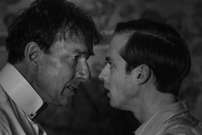 Black and white still of an older white man looking bedraggled and a more put together younger white man facing off